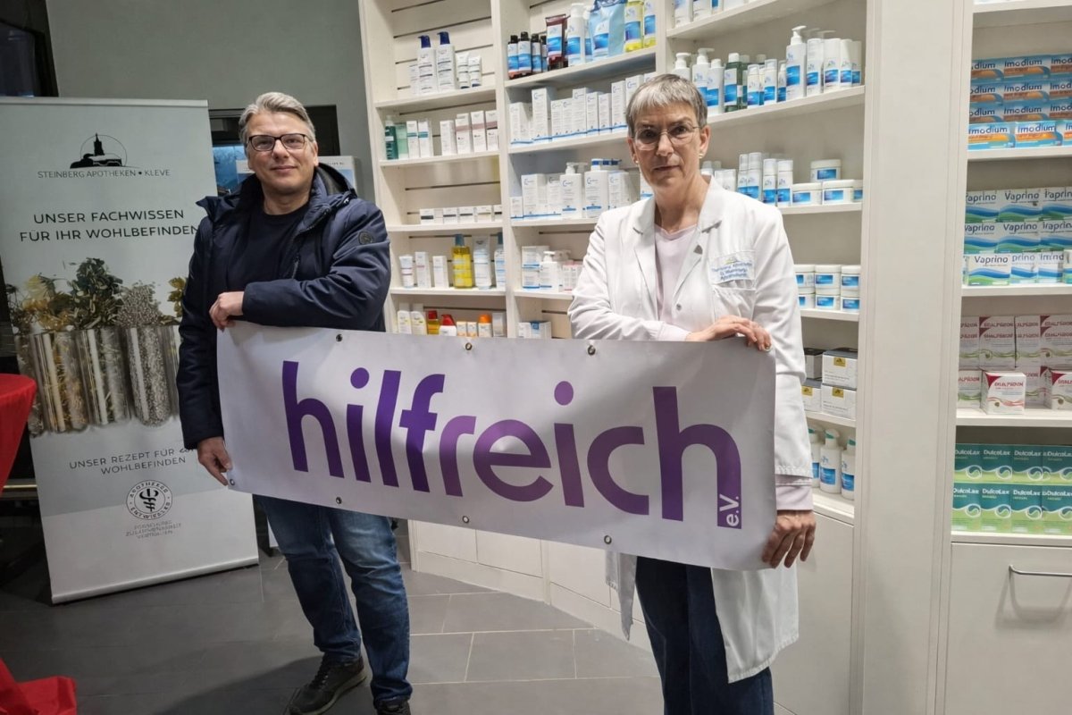 Beate Steinberg und Steffen Thewis halten gemeinsam ein Banner des Vereins hilfreich e.V. in einer Apotheke.